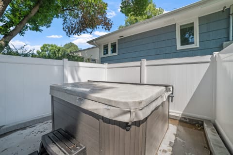 Hot tub in a backyard enclosed by a white vinyl fence, with green trees and a blue house in the background.