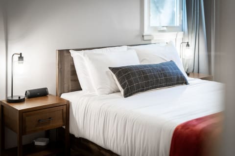 A well-decorated bedroom with white bedding, a plaid accent pillow, a bedside lamp, and a speaker on a nightstand.