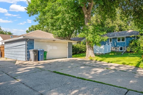 A light blue garage next to a driveway with trash bins and lush greenery.