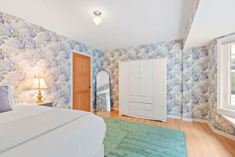 A bright bedroom with floral wallpaper, a white bed, and a wardrobe.