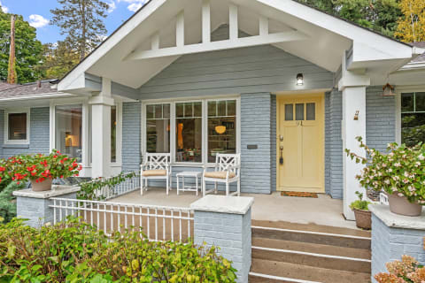 The front porch of a cozy house featuring two white chairs, a yellow door, and colorful flower pots.