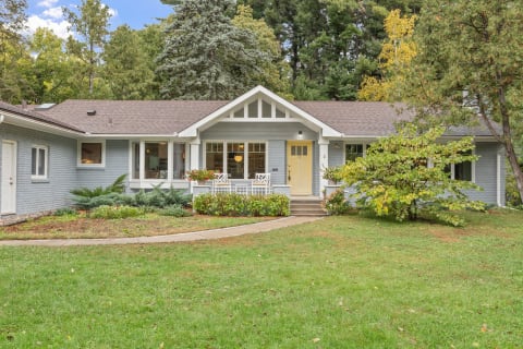 A single-story house with gray siding and a yellow front door, surrounded by greenery.