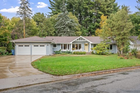 A light blue single-story house with a front porch and two-car garage, surrounded by trees and grass.