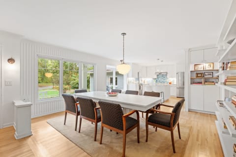 A spacious dining area with a long white table and dark upholstered chairs, featuring large windows and a modern kitchen in the background.