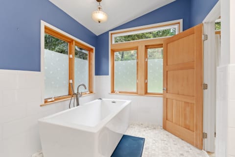 A modern bathroom featuring a white bathtub, blue walls, and large windows with a natural view.