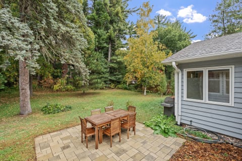 Backyard patio with a wooden table and chairs, surrounded by trees showing autumn foliage.