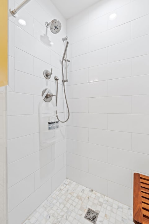 A clean and modern shower area with white tiles and a wooden bench.