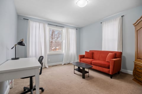 A cozy living room featuring an orange sofa, desk, and large windows with white curtains.