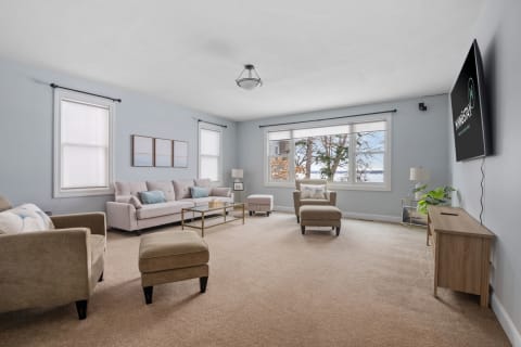 A spacious living room featuring light blue walls, a light-colored sofa, armchairs, large windows, and a flat-screen TV.