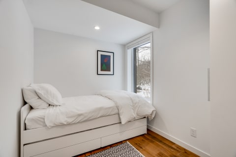 Minimalist bedroom featuring a single bed, white bedding, and a snowy view through a large window.