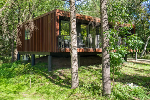 A contemporary brown cabin elevated on stilts in a wooded area with a deck and chairs.