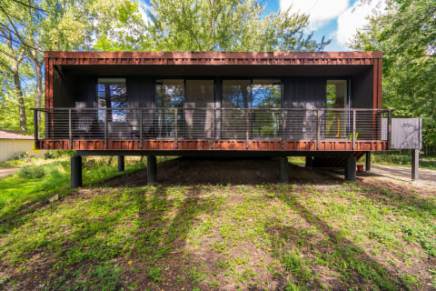 Contemporary house on stilts with a wooden and metal facade, featuring a spacious deck and large windows, surrounded by trees.