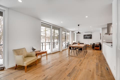 Modern open-concept living area with a dining table and armchair, showcasing large windows and wooden flooring.