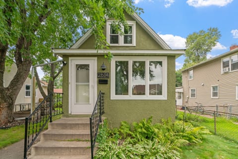 Small green house with a sloped roof, large windows, and a welcoming entrance in a garden setting.