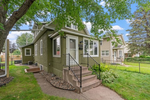 A two-story house with a green exterior, white doors, and windows, featuring a stone pathway and lawn.