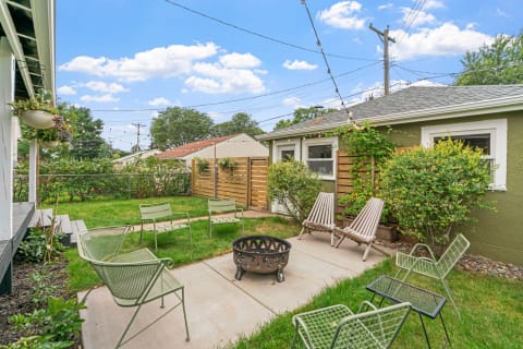 Cozy yard with a fire pit, green chairs, and greenery under a blue sky and clouds.