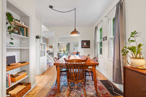 A dining room with a wooden table, chairs, and a pendant light, surrounded by natural light and decor.