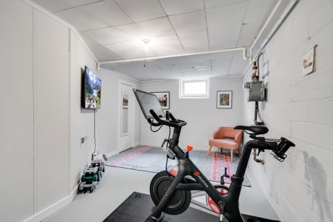 A basement home gym with a Peloton bike, TV showing a mountain landscape, and a comfortable chair.