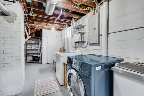 Interior view of a basement laundry room featuring a blue washing machine, a utility sink, and shelving for supplies.