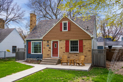 Charming home with a brick facade and red shutters, featuring adirondack chairs in the front yard.