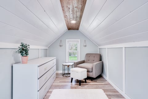 Cozy attic interior with a gray armchair, ottoman, and white dresser under sloped ceiling.