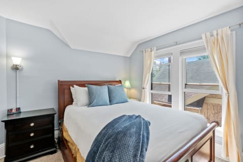 A cozy bedroom featuring a wooden bed, blue pillows, and large windows with cream curtains.