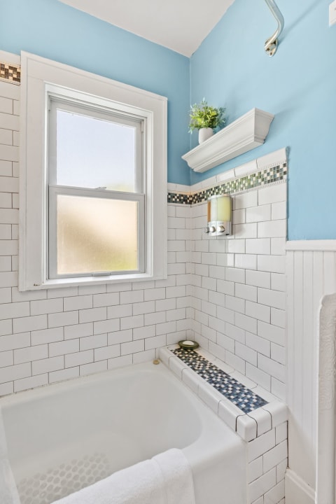 A bathroom corner with a white bathtub, tiled walls, a window, and a small plant on a shelf.