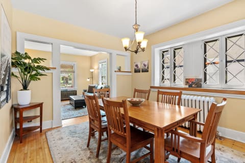 Bright dining area with wooden table, chairs, and potted plant, leading into a living room.