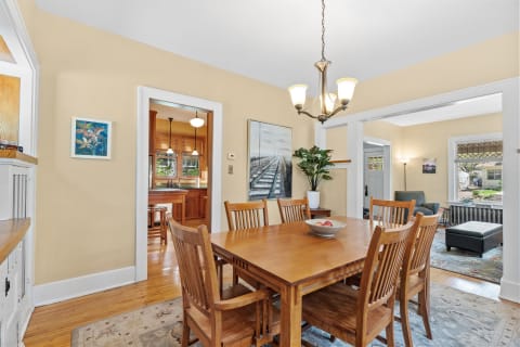 A cozy dining room connected to a kitchen, showcasing a wooden dining table, chandelier, and artwork.