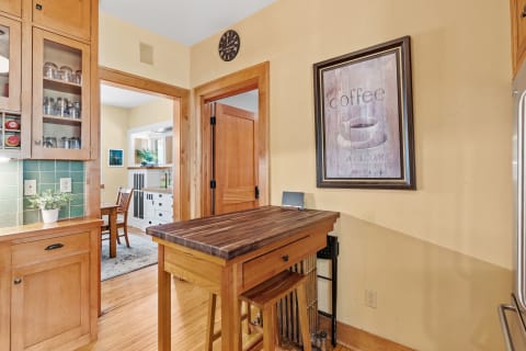 Interior view of a warm kitchen featuring a butcher block island, coffee-themed artwork, and glass-front cabinets.