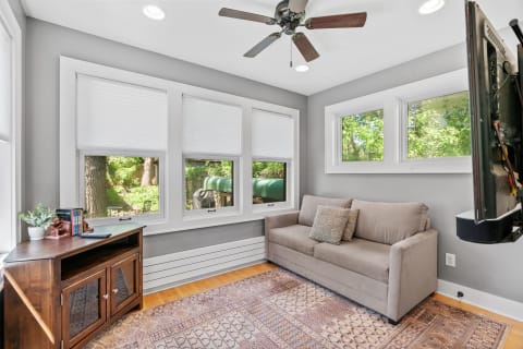 A bright sunroom featuring a gray sofa, wooden furniture, and large windows with greenery outside.