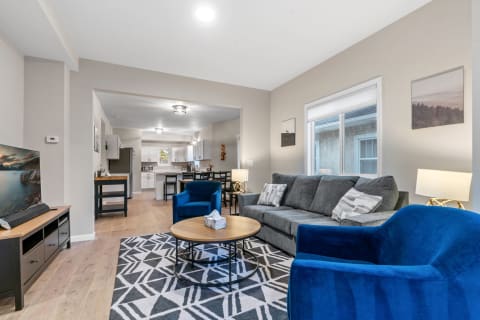 Bright living room featuring a gray sofa, blue chairs, and an open kitchen in the background.