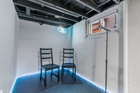 A basement corner with two chairs, a microphone, and a neon live music sign.