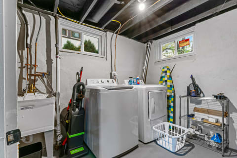 Laundry room with washing machine, dryer, ironing board, and storage unit in a basement setting.