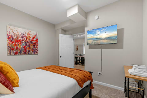 A neatly styled bedroom featuring a bed with an orange blanket, decorative pillows, a wall-mounted TV, and a piece of floral wall art.
