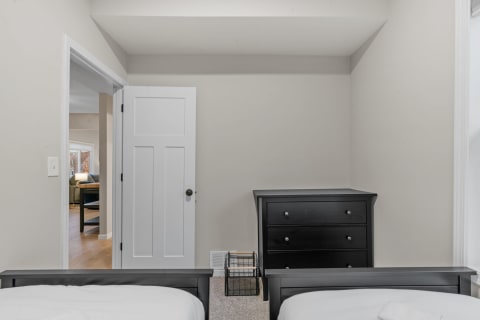 Minimalist bedroom featuring twin beds and a dresser with an adjoining room in the background.