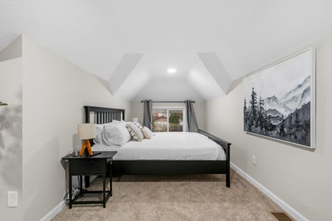 A cozy attic bedroom featuring a sloped ceiling, double bed, nightstand with lamp, and mountain artwork.