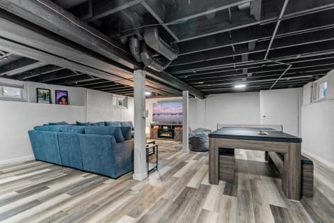 A modern basement transformed into a cozy recreation room with a sectional couch, game table, and a TV wall.