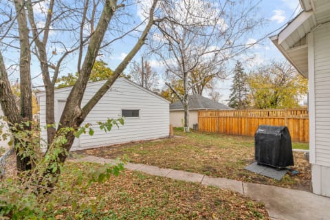 Outdoor backyard view with a white shed and a covered grill surrounded by trees and fallen leaves.