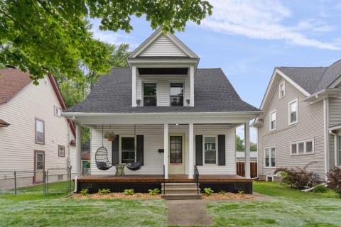 A white house with black shutters and a spacious porch featuring hanging chairs and a small table, surrounded by lush green grass.