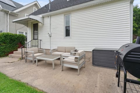 Outdoor seating set with beige cushions and a grill by a white house.