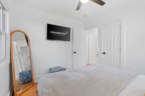 A cozy bedroom with a bed, a full-length mirror, a flat-screen TV, and white walls.