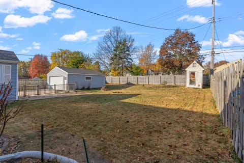 A fenced backyard with autumn trees, a gray garage, and a small shed under a blue sky.