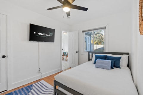 Modern bedroom with gray metal bed frame, blue decorative pillows, wall-mounted TV, and natural light from a window.