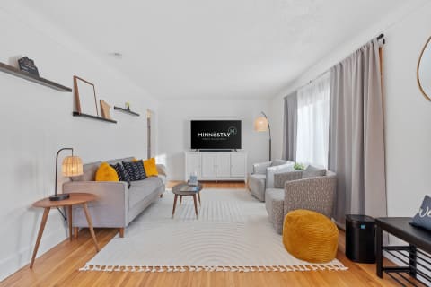 A modern living room with gray sofas, yellow pillows, a circular rug, and a television displaying MINNESTAY.