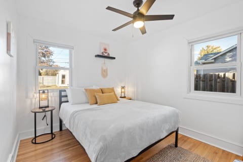 Cozy bedroom with white walls, wooden floor, a bed with white and mustard pillows, and natural light from two windows.