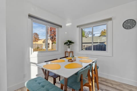 Bright dining space with a white table and teal chairs, featuring yellow placemats and natural light from two windows.