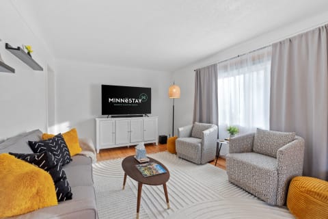 Modern living room with gray couch, patterned chairs, and bright yellow pillows, featuring a TV and natural light.