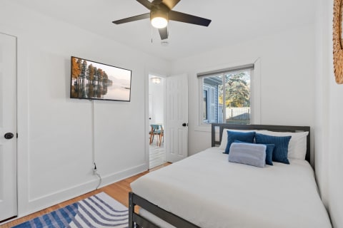 A modern bedroom with a bed, decorative pillows, a wall-mounted TV, and a ceiling fan, bathed in natural light.