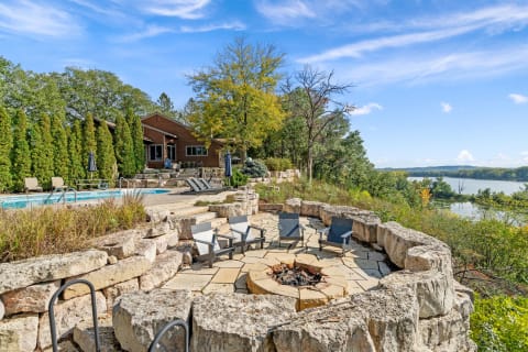 Outdoor area with a fire pit, pool, and wooden house amidst trees and sky.